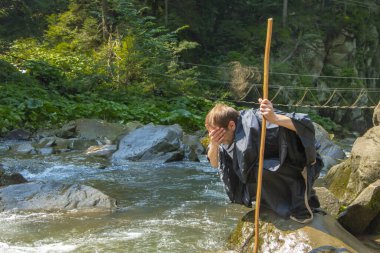 45-50 yaşındaki siyah cüppeli bir keşiş ve bir sopayla yüzünü bir dağ nehrinden gelen suyla yıkar şelaleli kayalık bir dağ manzarasının arka planına karşı.,