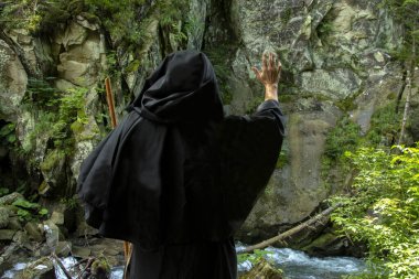 45-50 yaşlarındaki yaşlı bir keşişin portresi, siyah cüppeli ve asalı bir sakallı, bir dağ şelalesinin ve ormanın arka planında seçici bir odak noktası var. Konsept: Büyü ve büyü, bir taştan akan su..