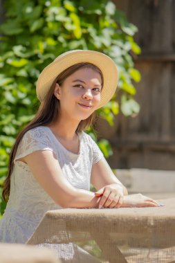 17-20 yaşlarında, hasır şapkalı ve beyaz elbiseli genç bir kız, ahşap bir masada yeşil üzümlerden ve eski bir kapıdan oluşan bir arka planda oturuyor..