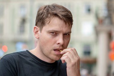 Bulanık bir şehrin arka planında patates kızartması yiyen genç bir adamın sokak portresi. Konsept: Fast food sokak yemekleri, şehirde hafif atıştırmalıklar.