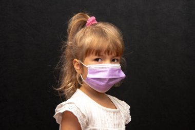 Siyah arka planda sarı saçlı, maske takmış küçük bir kızın stüdyo portresi..