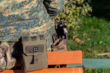 Kamuflajlı bir asker dinleniyor, çantalar ve kılıflar yakında görünüyor.