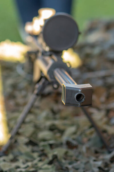 Снайперская винтовка с оптическим прицелом установлена на двуногих и лежит на камуфляжном покрытии.