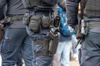 Polis memurları taktik ekipman ve silah kılıflarıyla caddenin yakınında duruyorlar.