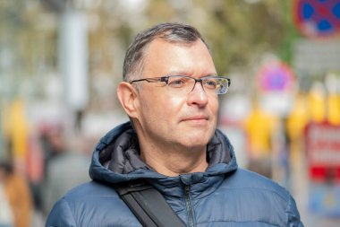 Mavi ceketli 50-55 yaşındaki bir adam bulanık bir şehir geçmişinin önünde poz veriyor.