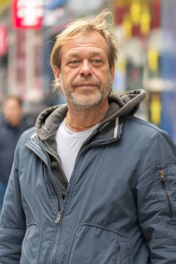 Sakallı yaşlı bir adam yayaların arka planına karşı bir şehir caddesinde duruyor.