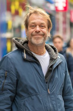 50-55 yaşındaki ceketli bir adam, kalabalık bir caddede yoldan geçenlerin arasında dururken gülümsüyor.