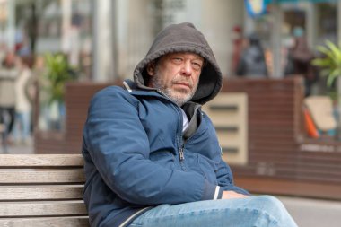 Kalın elbiseli bir adam şehir caddesinde bir bankta oturuyor..