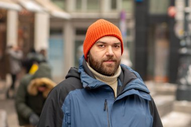 Kış kıyafetleri içinde sakallı bir adam bulanık bir şehir geçmişine sahip bir sokakta duruyor.