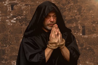 Siyah başlıklı yaşlı bir keşiş elleri bağlı ve tuğla bir duvara karşı tespihle dua ediyor.