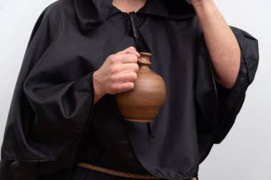 Siyah cüppeli keşişin elleri beyaz arka plandaki stüdyoda izole edilmiş kilden sürahi taşıyor.