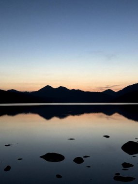 Sakin suda yansıtılan sessiz dağ çizgisi, sakin göle yansıyan siluetli dağların üzerinde yumuşak eğimli gökyüzü.