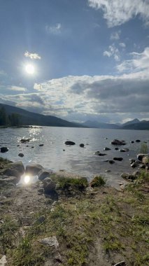 Parlayan yansımalarıyla dramatik göller, engebeli göl kıyısında dağ siluetiyle canlı günbatımı yansımaları.