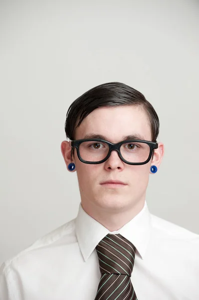 Молодой человек с очками и проколотыми ушами, одетый в белую рубашку и коричневый галстук перед камерой Лицензионные Стоковые Фото