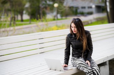 Girl works with laptop computer sitting on a bench in the park looking to camera and smiling