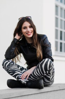 Beautiful young female student in jacket sitting down on an old stone wall outdoors and smiling during autumn spring day