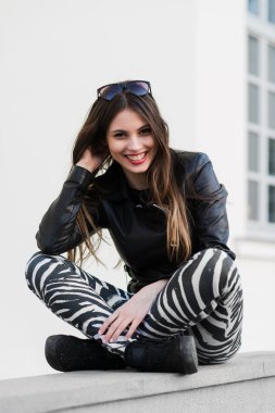Beautiful young female student in jacket sitting down on an old stone wall outdoors and smiling during autumn spring day