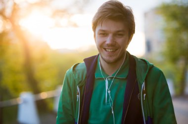 Genç adam kulak kulaklık ile müzik dinlemek şehir etrafında yürüyüş kameranın arka ışık ve gülümseyerek bakar. Yakın çekim shot. Gün batımı. Sonbahar hava.