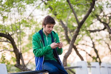 Gün batımı dokunaklı cep telefonu müzik parkta oturan genç erkek öğrenci. Yumuşak ışık ve vintage renk