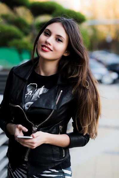Chica caminando y mensajes de texto en el teléfono inteligente en la calle con una chaqueta de cuero en verano — Foto de Stock