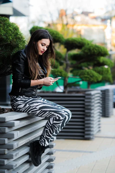 Schöne junge dunkelhaarige Frau schreibt auf ihrem Smartphone SMS, während sie auf der Bank im Stadtpark sitzt — Stockfoto