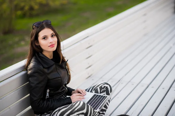 Portræt af en glad kvindelig studerende sidder på bænken med laptop udendørs og ser på kameraet - Stock-foto