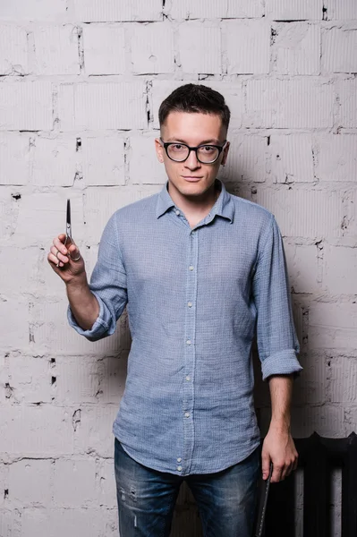 Портрет молодого чоловіка-перукаря в окулярах, позує з ножицями, на сірому цегляному тлі стіни, вид спереду, вертикальне зображення — стокове фото