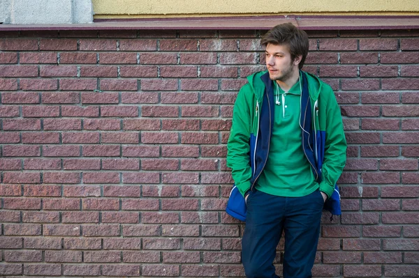 Gewone jonge man dragen groene t-shirt jas en jeans tegen een rode bakstenen muur zijwaarts op zoek — Stockfoto