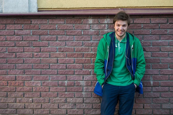 Joven común con chaqueta de camiseta verde y jeans contra una pared de ladrillo rojo mirando a la cámara — Foto de Stock