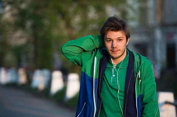 Young man walking around city listening to music with ear headphones looks at the camera and smiling. Close-up shot. Sunset. Autumn weather. — Stock Photo, Image