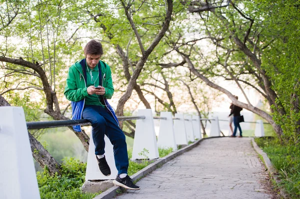 Joven estudiante masculino sentado en el parque al atardecer tocando el teléfono móvil escuchando música. Luz suave y color vintage — Foto de Stock