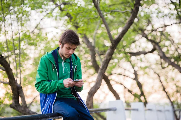 Jonge mannelijke student zitten in het park bij zonsondergang ontroerende mobiele telefoon luisteren naar muziek. Zacht licht en vintage kleur — Stockfoto