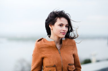 Yakın çekim portre deri ceket gülümseyen ve arka plan ışığı açık havada duran giyen genç mutlu güzel çekici kadın. Rüzgar saçlarını havaya.