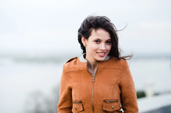 Primer plano retrato de la joven hermosa mujer atractiva feliz con abrigo de cuero sonriendo y de pie sobre la luz al aire libre de fondo. El viento sopla su pelo . — Foto de Stock