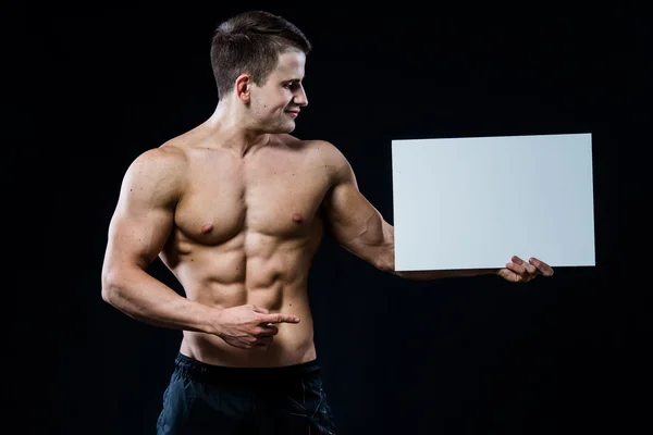Shirtless muscular young man standing holding a blank horizontal white banner pointing finger at the copyspace for your text, on black — Stock Photo, Image