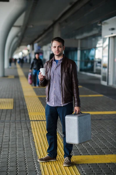 Молодой привлекательный мужчина пьет кофе по пути к терминалу аэропорта, глядя в камеру — стоковое фото