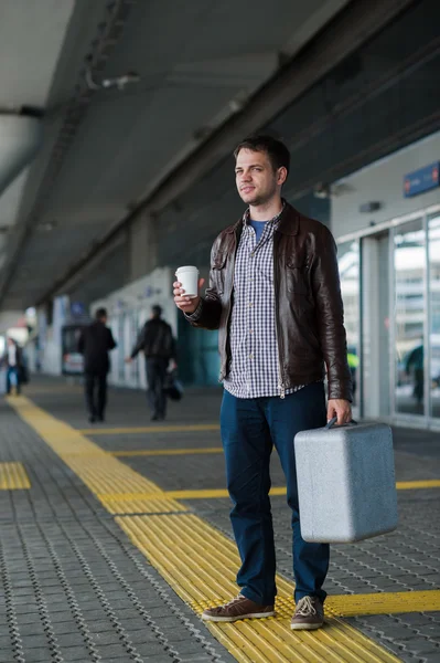 Full längd porträtt unga glada leende stilig resande mannen i 20-talet lämnar ankomster flygplatser lounge terminal byggnaden efter att samla in sitt bagage med en kopp nybryggt kaffe — Stockfoto