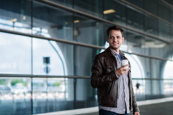 Elegante joven viajero masculino guapo con cerdas de pie al aire libre cerca de la terminal del aeropuerto. Hombre con chaqueta y camisa. Sonriente persona mirando a la cámara sosteniendo la taza de café —  Fotos de Stock