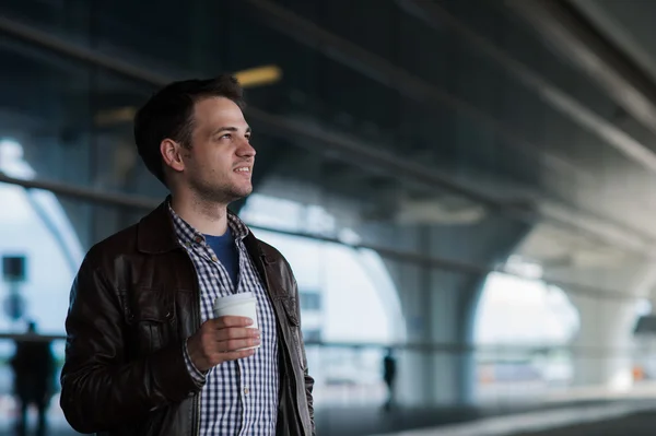 Stylish handsome young male traveller with bristle standing outdoors near the airport terminal. Man wearing jacket and shirt. Smiling person looking to camera holding cup of coffee — Stock Photo, Image