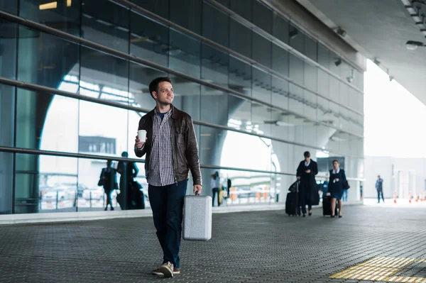 Портрет молодого человека, идущего рядом с аэропортом с чемоданом и кофе — стоковое фото
