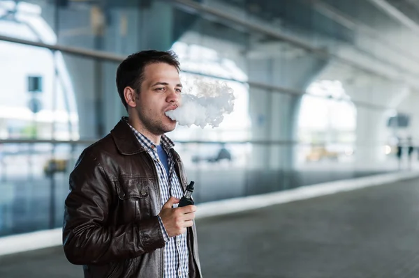 Man with a bristle smoking e-cigarette vaporizer box mode outdoors near the airport terminal before flight — Stock Photo, Image