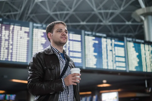 Ung man med en väska i flygplats nära flyg tidsplanen håller kopp kaffe — Stockfoto