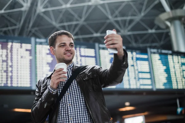 Glimlachend jonge reiziger man met een kopje koffie op de luchthaven maakt selfie voor tijdschema bestuur — Stockfoto