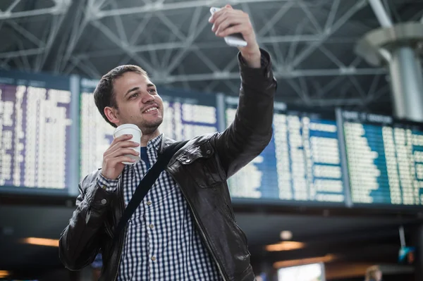 Glimlachend jonge reiziger man met een kopje koffie op de luchthaven maakt selfie voor tijdschema bestuur — Stockfoto
