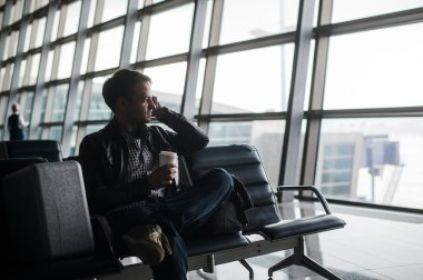 Modern havaalanında smartphone kullanarak bankta oturmuş rahat tarzı elbise giyen genç yakışıklı adam portresi. Bagaj ile seyahat eden yolcu uçağı için bekleme yapma çağrısı, çanta