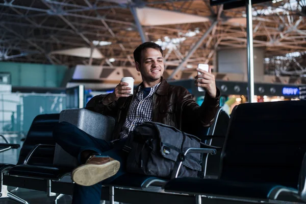 Porträtt av en ung man skrattar och tar selfie med kaffe på flygplatsen väntar på sin flight — Stockfoto