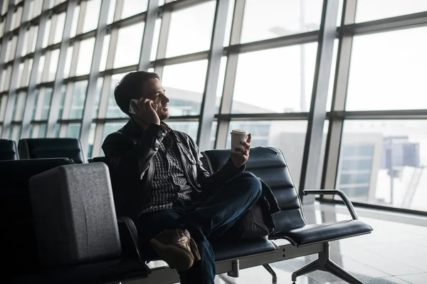 Portrait of young handsome man wearing casual style clothes sitting on the bench in modern airport using smartphone. Passenger travelling with luggage bag making call, while waiting for his flight — Stock Photo, Image
