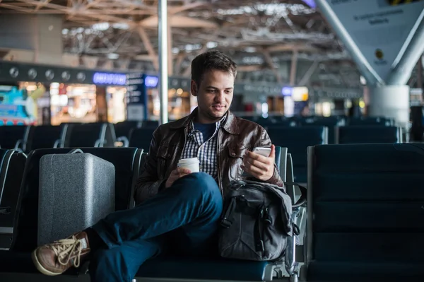 Красивый улыбающийся мужчина в повседневной одежде, держащий багаж и отправляющий сообщения через свой мобильный телефон, сидя в зале аэропорта с чашкой кофе — стоковое фото
