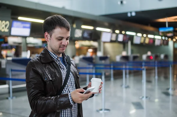 Retrato de homem bonito jovem andando no terminal do aeroporto moderno, mensagens de texto, viajar com saco e café, vestindo roupas de estilo casual, mesas de registro borradas no fundo — Fotografia de Stock