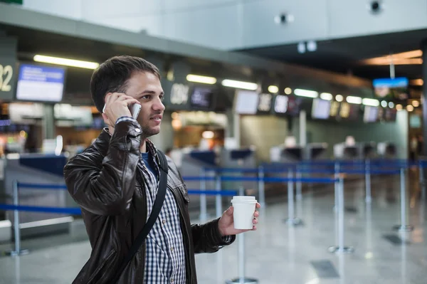 Porträtt av ung vacker man vandrar i modern flygplats terminal, talar smart telefon, reser med väska och kaffe, bär casual stil kläder och suddig registrering skrivbord på bakgrund — Stockfoto